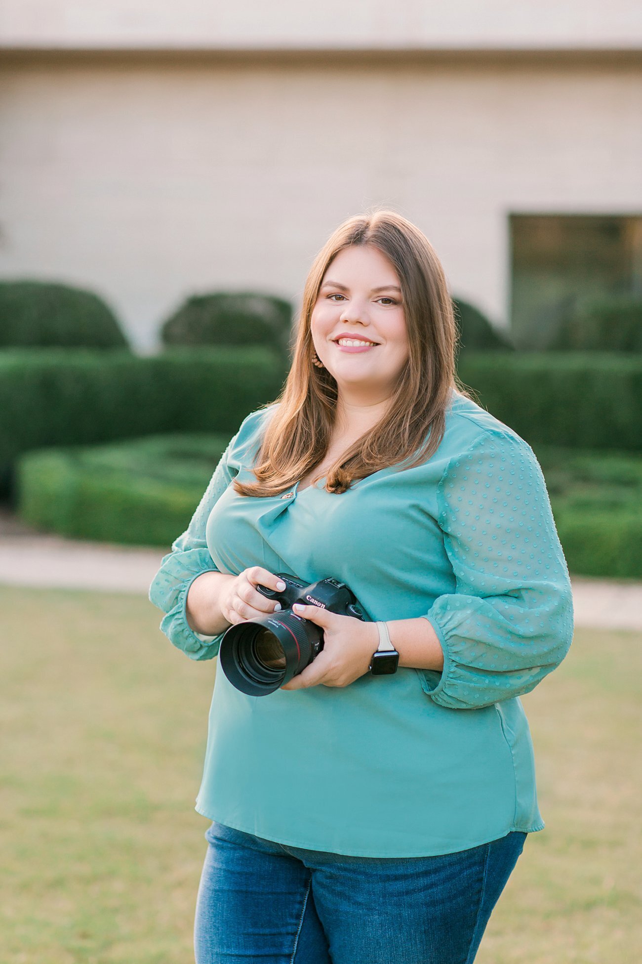Amanda Horne standing on a lawn in front of trimmed hedges, holding her camera and smiling at the lens.