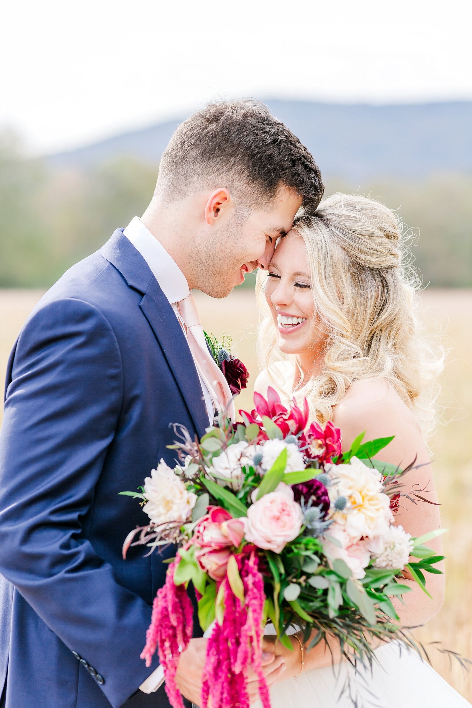 A bride and groom rest their foreheads together, laughing, holding a burgundy and blush bouquet in an open field.